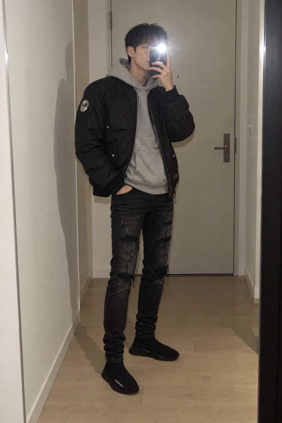 A Korean man takes a flash mirror picture in a hallway, wearing a black AAPE bomber over a grey Essentials hoodie, ripped Amiri jeans and Balenciaga Speed Trainers.