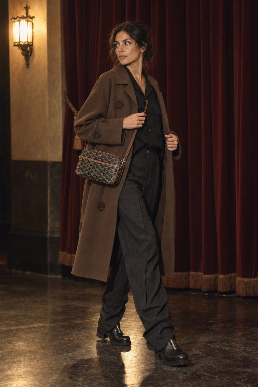 An Italian woman in a brown Song for the Mute coat, a black UJOH shirt and Avavav pinstripe trousers stands by a theatre's velvet curtain.