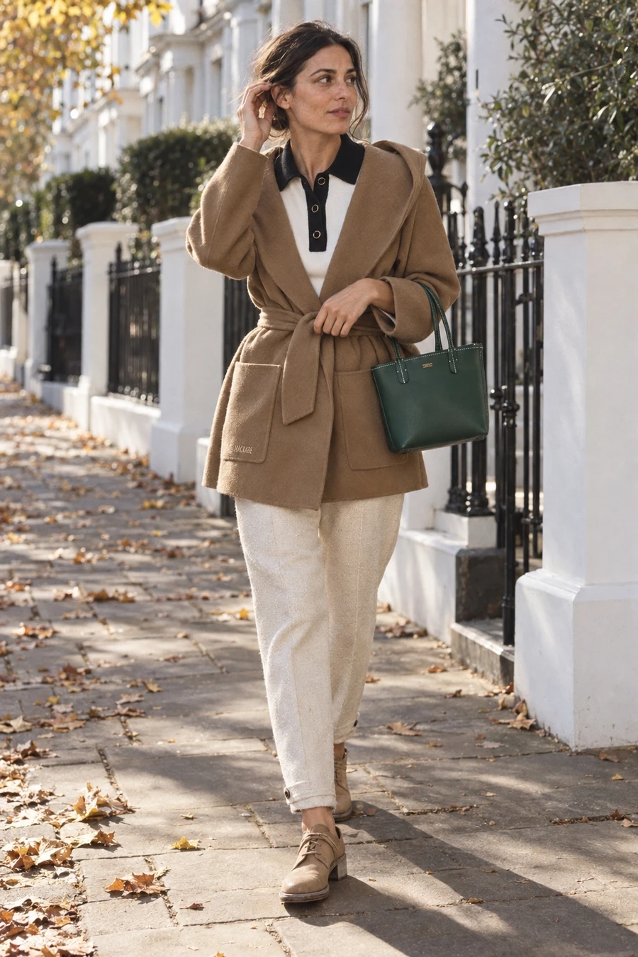 A woman walks past white townhouses on a sunny morning in a camel Mackage wrap jacket, a white Nina Ricci polo, ivory trousers and beige Guidi derbies.