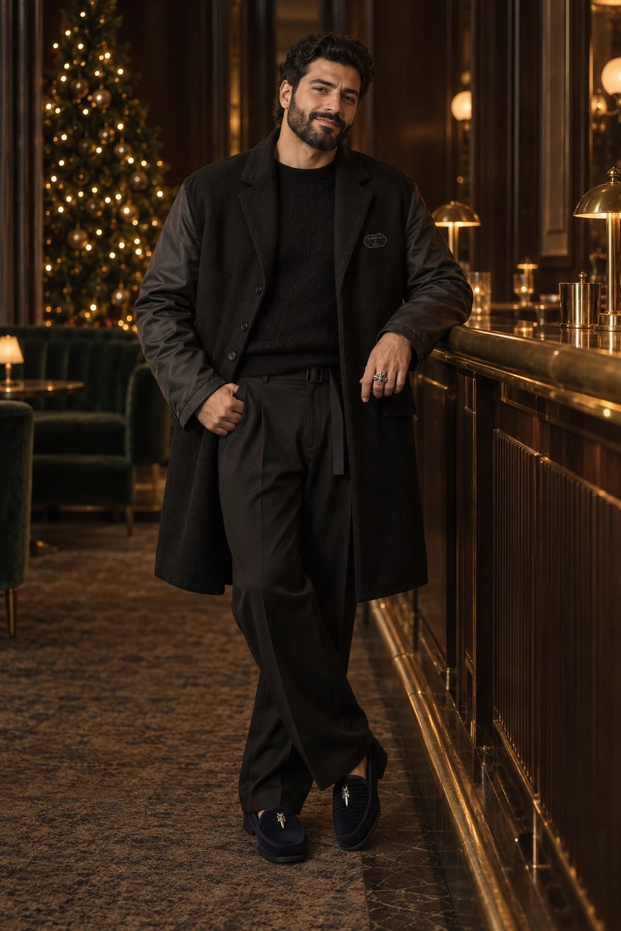 An Iraqi man in a black Undercover felted jacket, a Louis Vuitton knit, UJOH trousers and navy Chrome Hearts loafers stands at a hotel bar by a Christmas tree.