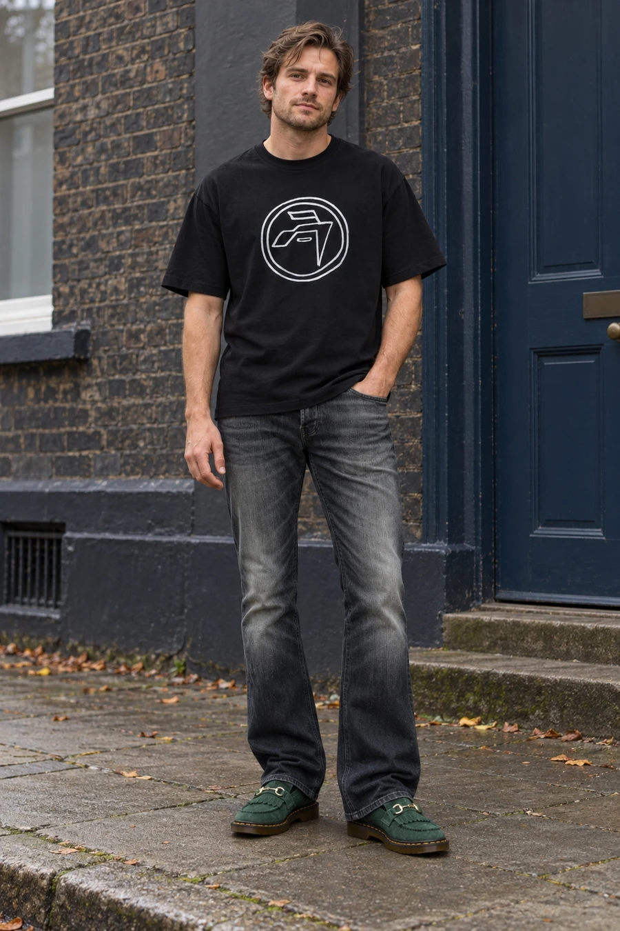 An adult man wears a black circle graphic tee, faded dark bootcut jeans and green suede loafers on a London street.