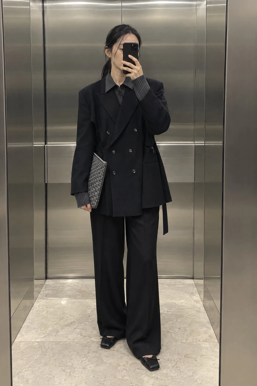 A woman photographs her reflection in an office lift mirror, wearing a black UJOH double breasted jacket, a Litkovska shirt, INTO THE EAST trousers and MM6 ballet flats.