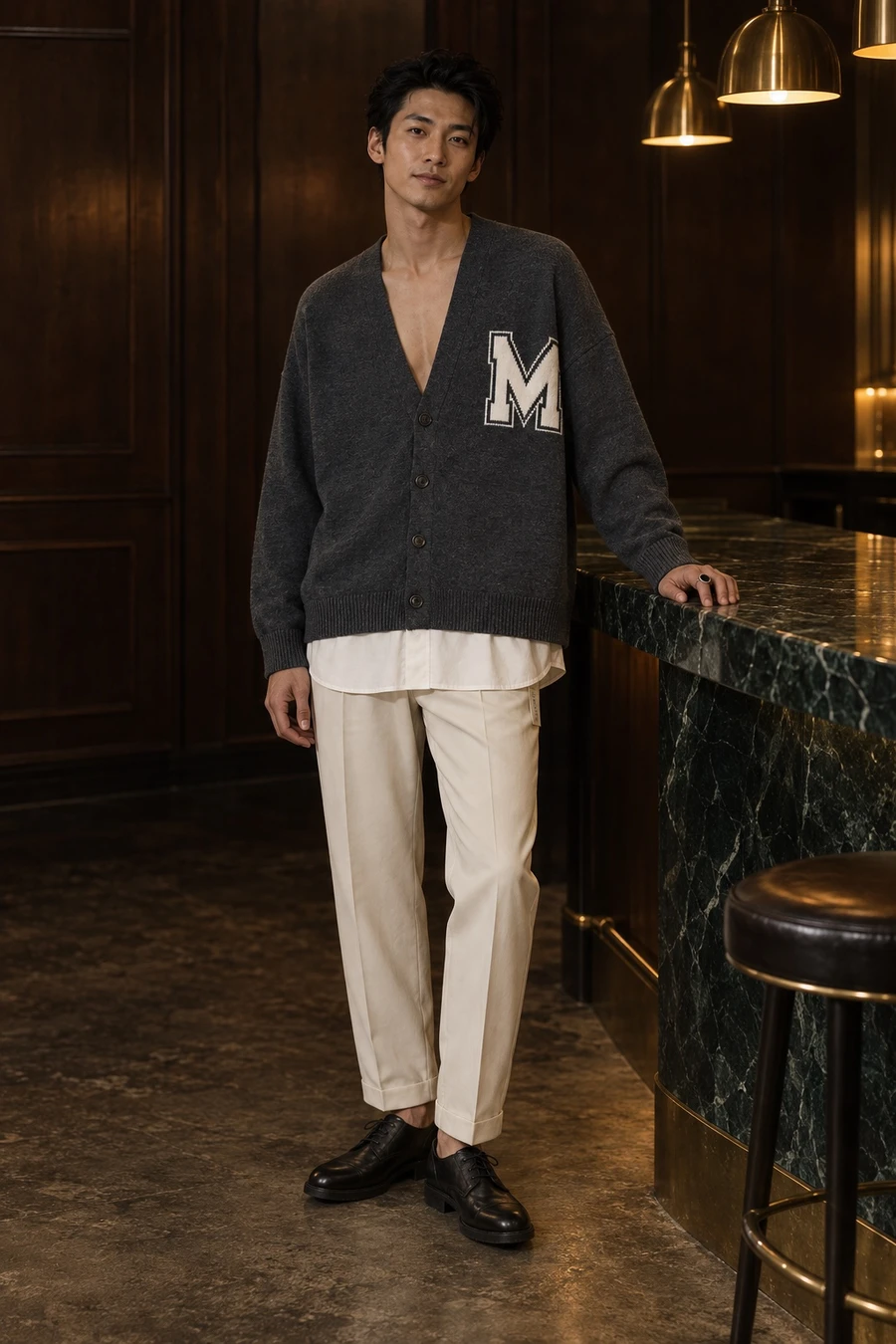 A man in a dark grey MM6 Maison Margiela cardigan, ivory Undercover trousers and black Guidi derbies stands at a marble bar at night.