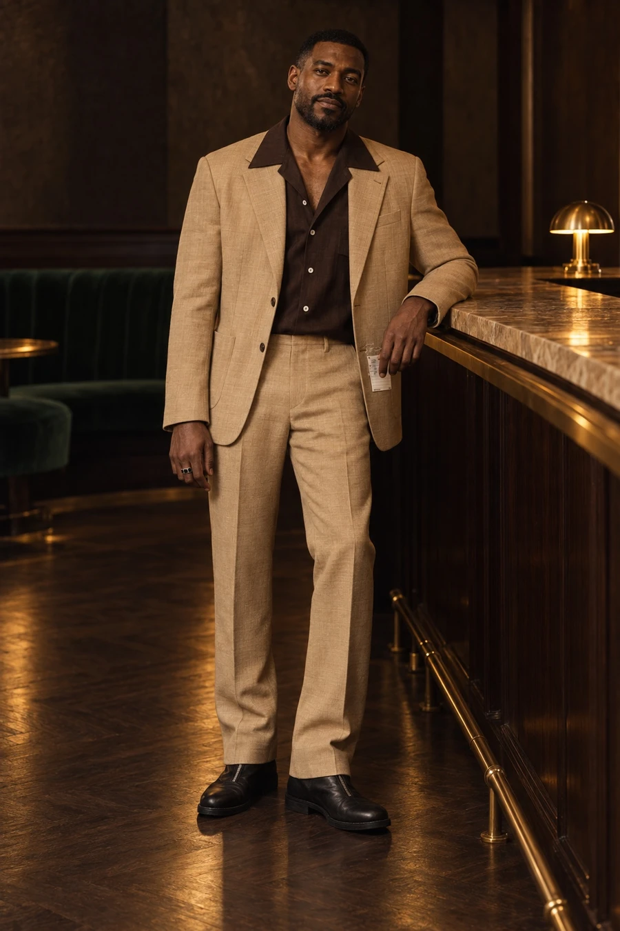 A man in an MM6 Maison Margiela camel suit, a chocolate UJOH camp collar shirt and black Guidi front zip boots stands at a hotel bar at night.
