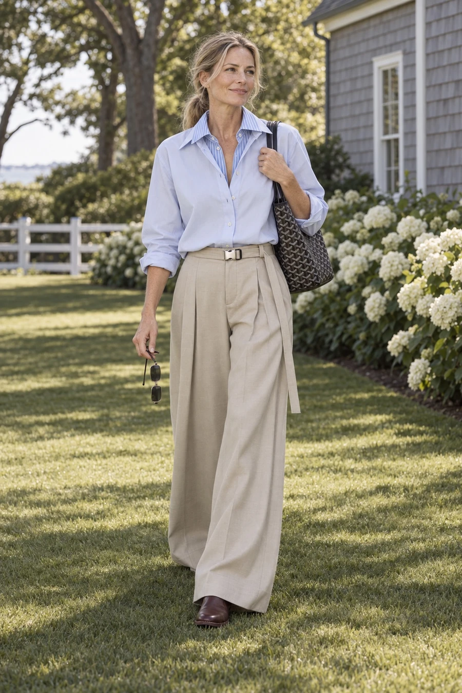 A woman in a pale blue Litkovska shirt and sand UJOH wide trousers carries a black Goyard Saint Louis tote across a lawn in the Hamptons.