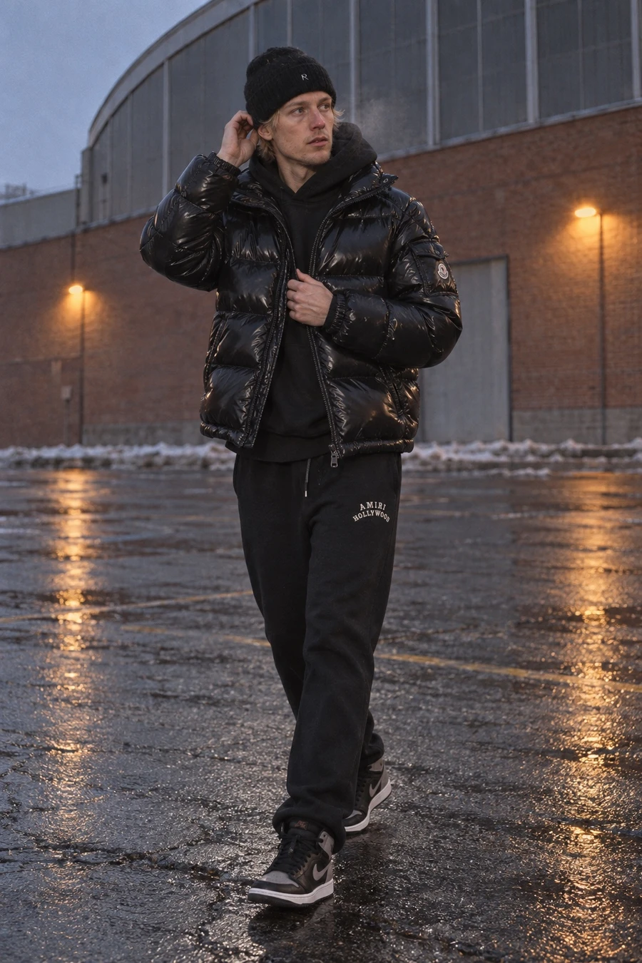 A Finnish man in a glossy black Moncler puffer, a black Essentials hoodie, Amiri sweatpants and Jordan 1 Shadows crosses an arena parking lot at dusk.