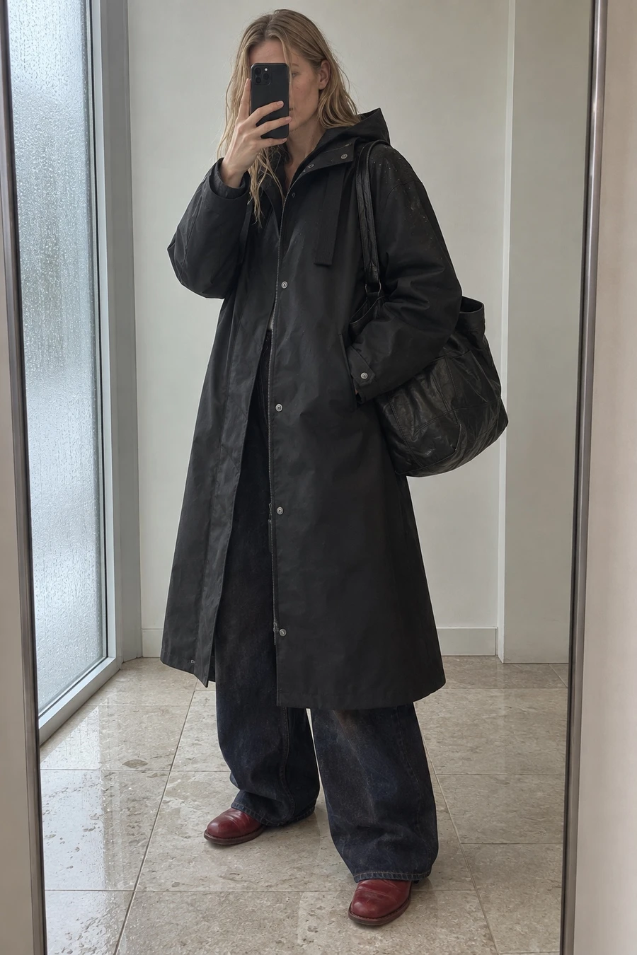 A woman photographs her reflection in a hallway mirror, wearing a long black g-lab parka, MM6 wide leg jeans and red Guidi boots, with a COOR tote.