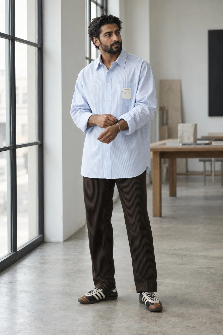 A man in an oversized MM6 Maison Margiela striped shirt, Homme Plissé chocolate pleated trousers and Wales Bonner Karintha sneakers stands by a studio window.
