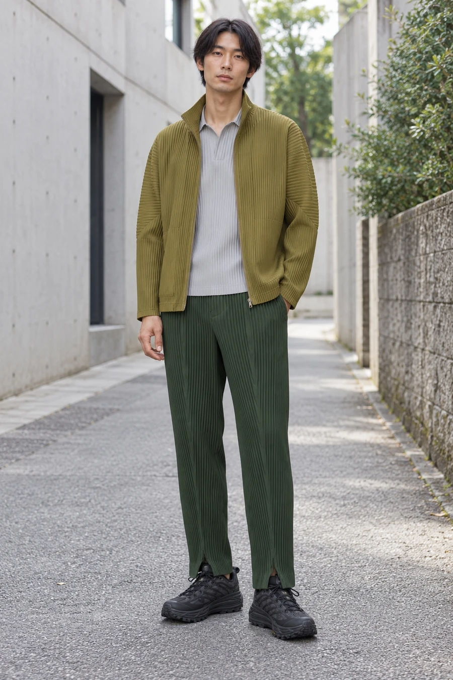 An adult man wears an olive pleated jacket, grey polo and dark green pleated trousers with black trail shoes in Tokyo.
