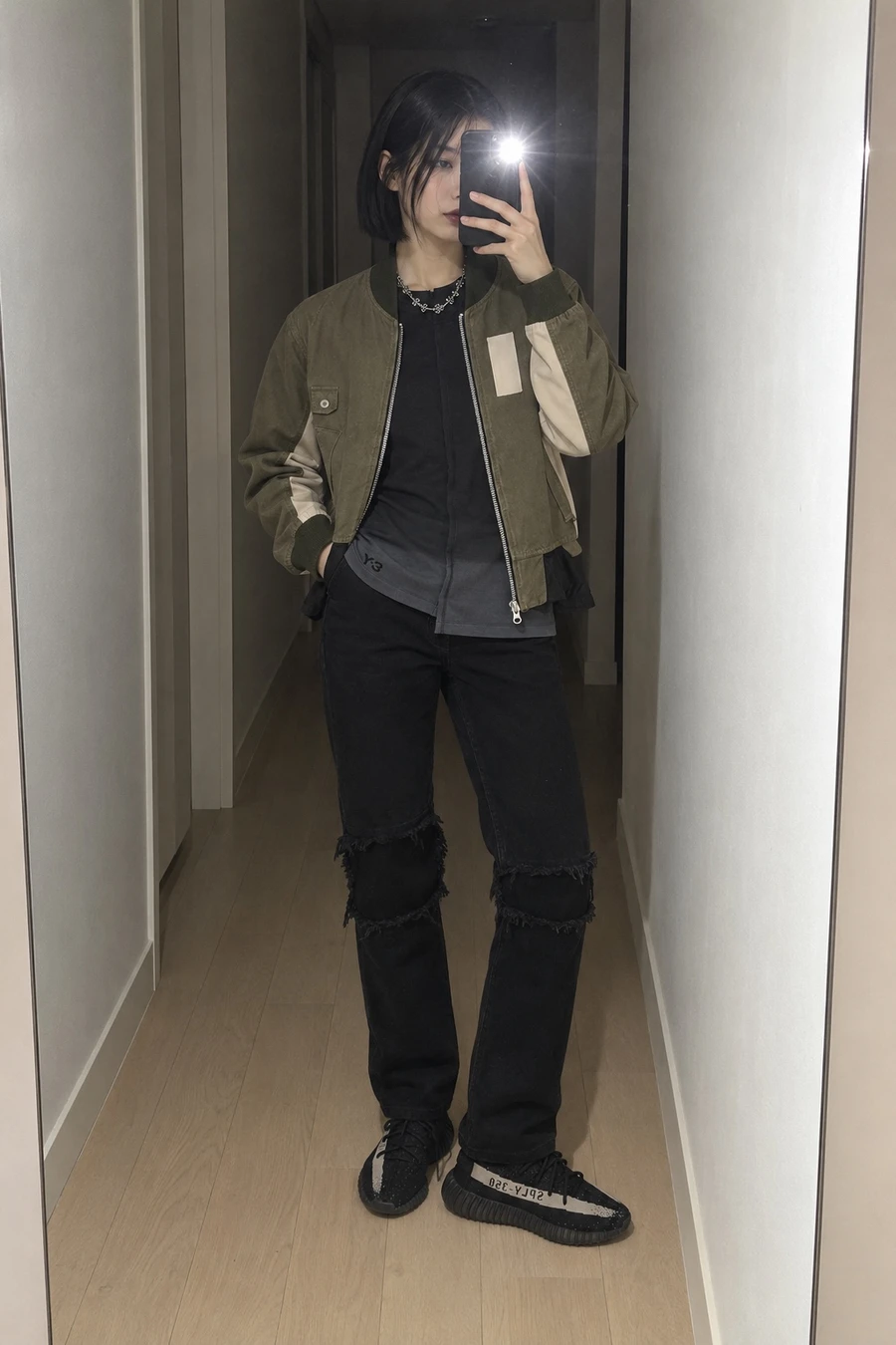 A woman takes a flash mirror photo in a hallway, wearing a green Song for the Mute bomber, a Y-3 tee, ripped black MM6 jeans and black sneakers.