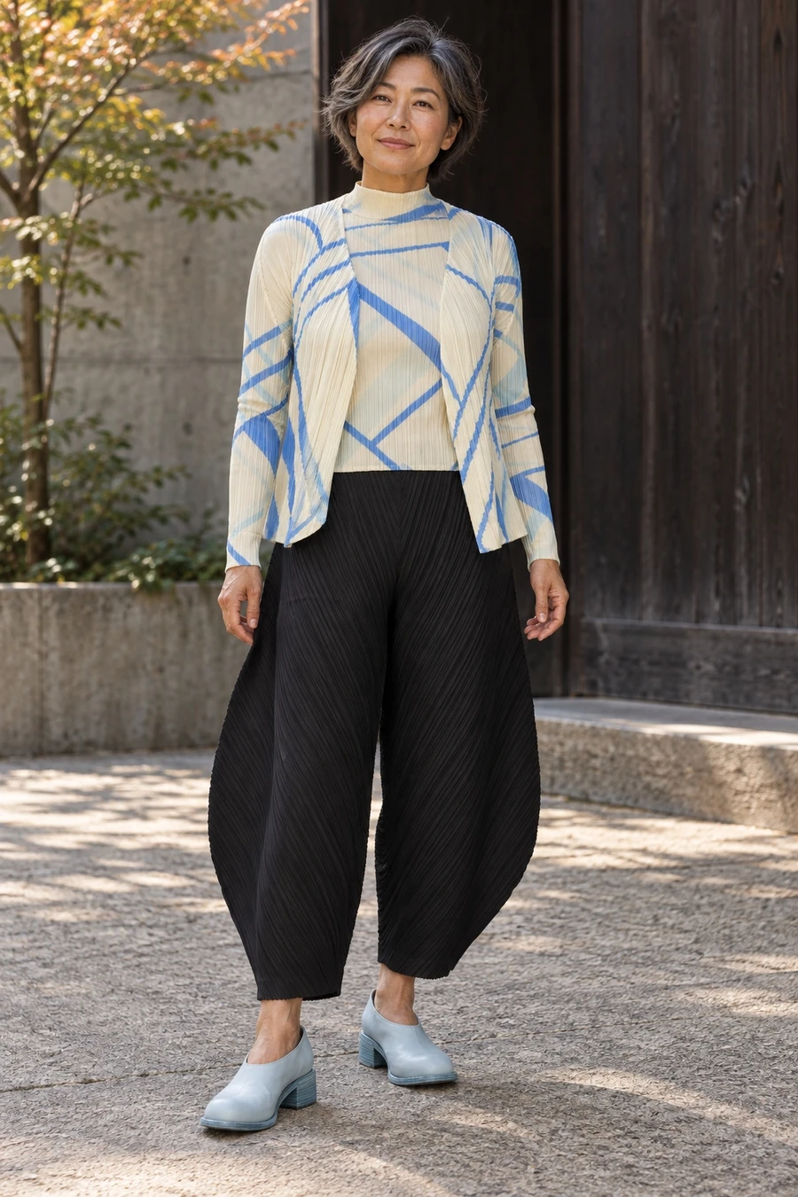 An adult woman wears cream and blue Pleats Please layers, black cocoon trousers and pale blue Guidi shoes in a Tokyo courtyard.