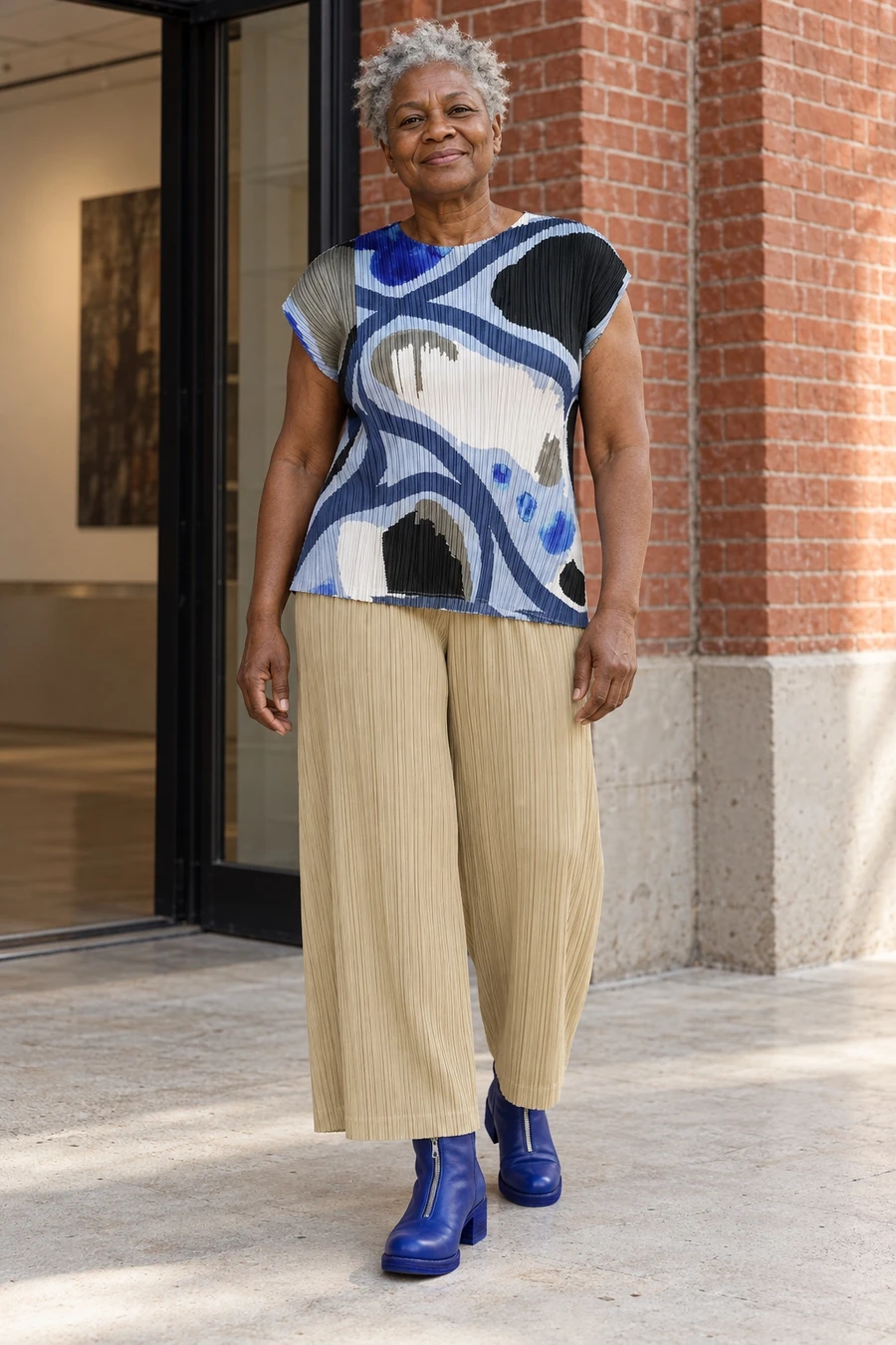 An adult woman wears a blue abstract Pleats Please top with beige pleated trousers and cobalt Guidi boots in Toronto.
