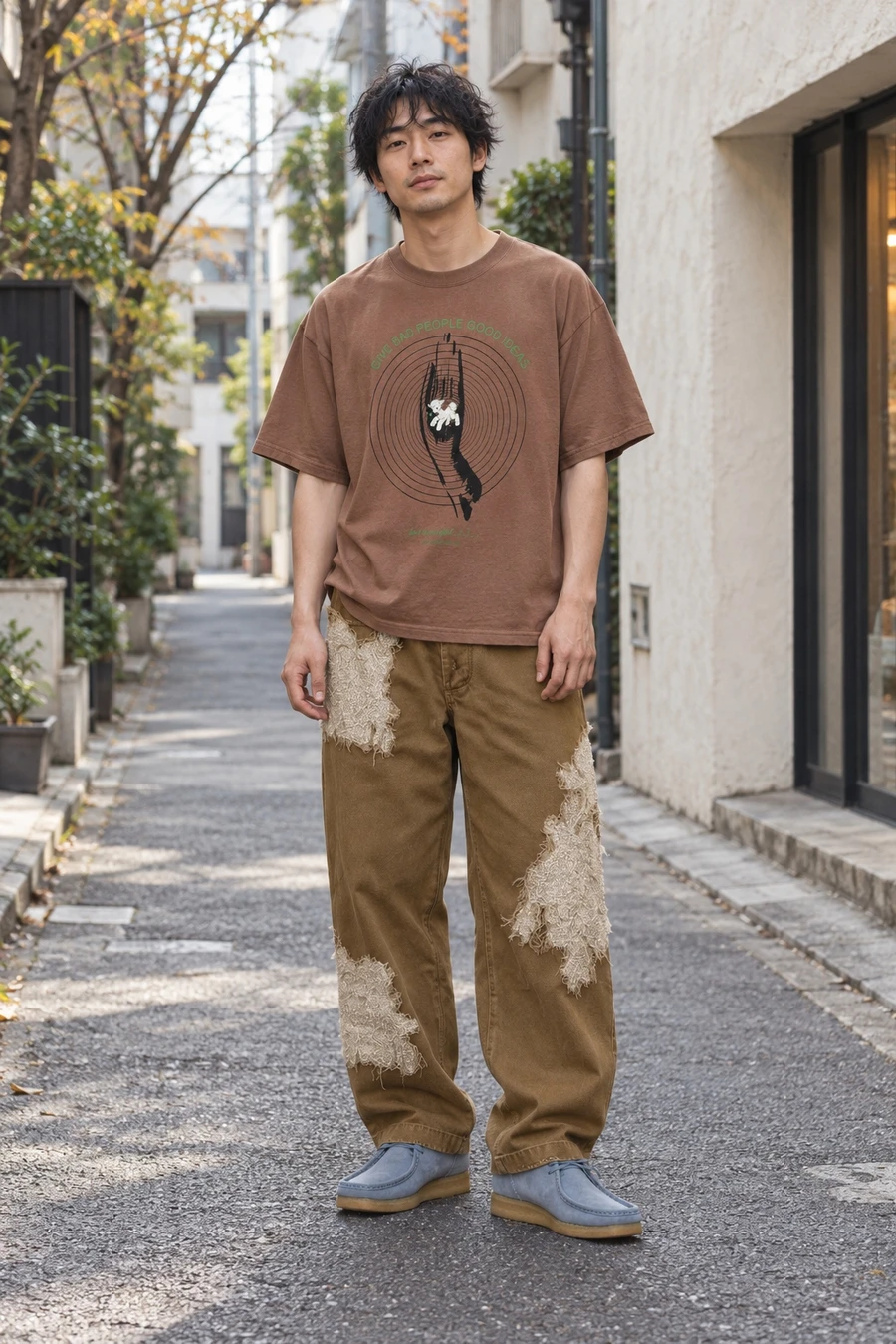 An adult man wears a brown graphic tee, khaki patchwork trousers and blue suede shoes on a Tokyo lane.