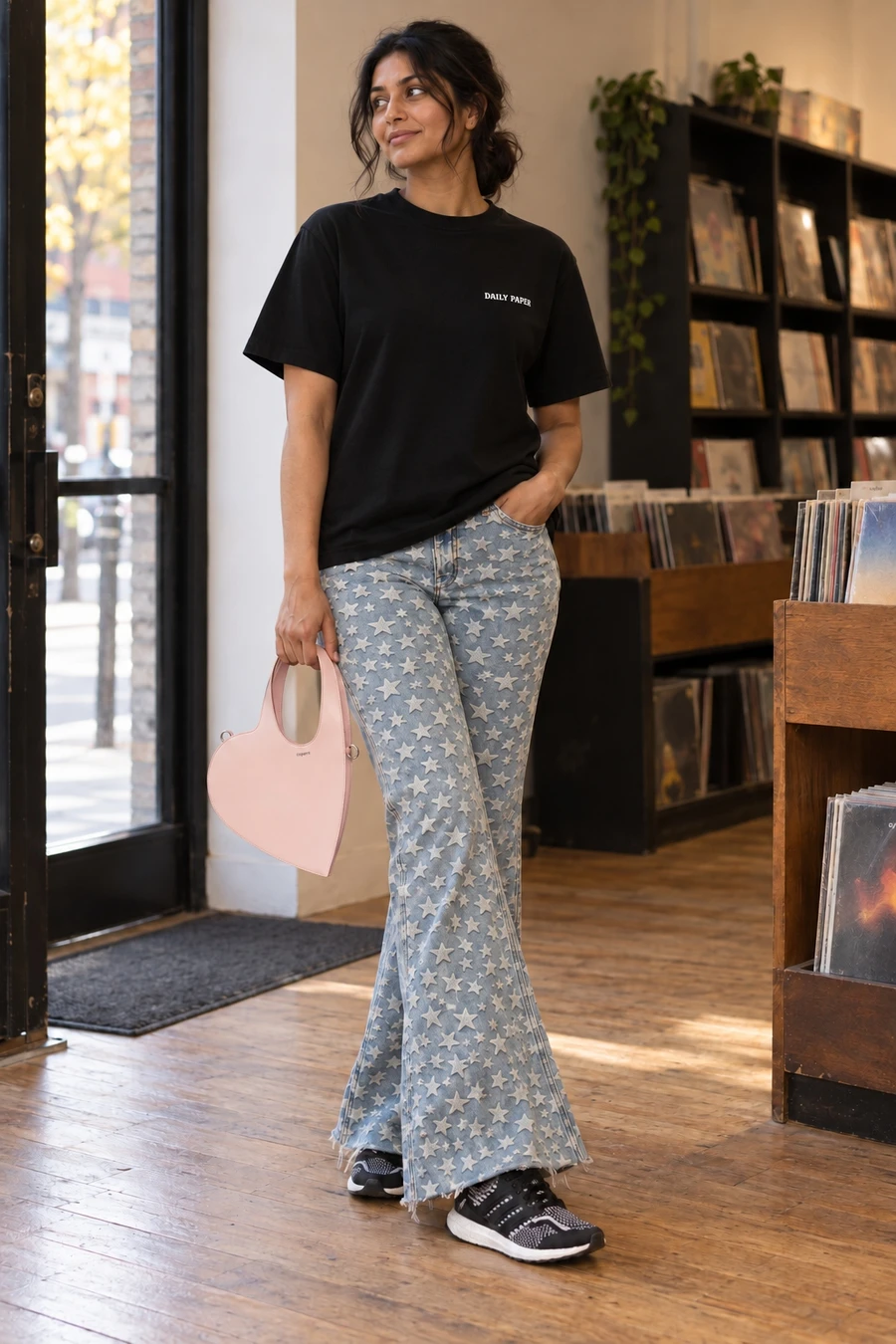 An adult woman wears a black Daily Paper tee, blue ERL star flares and black Adidas trainers, holding a pink Coperni heart bag in a Toronto doorway.