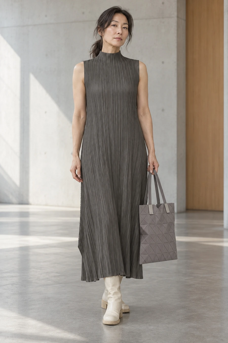 An adult woman wears a charcoal Pleats Please high neck dress with white Guidi boots and a grey Bao Bao tote in Tokyo.