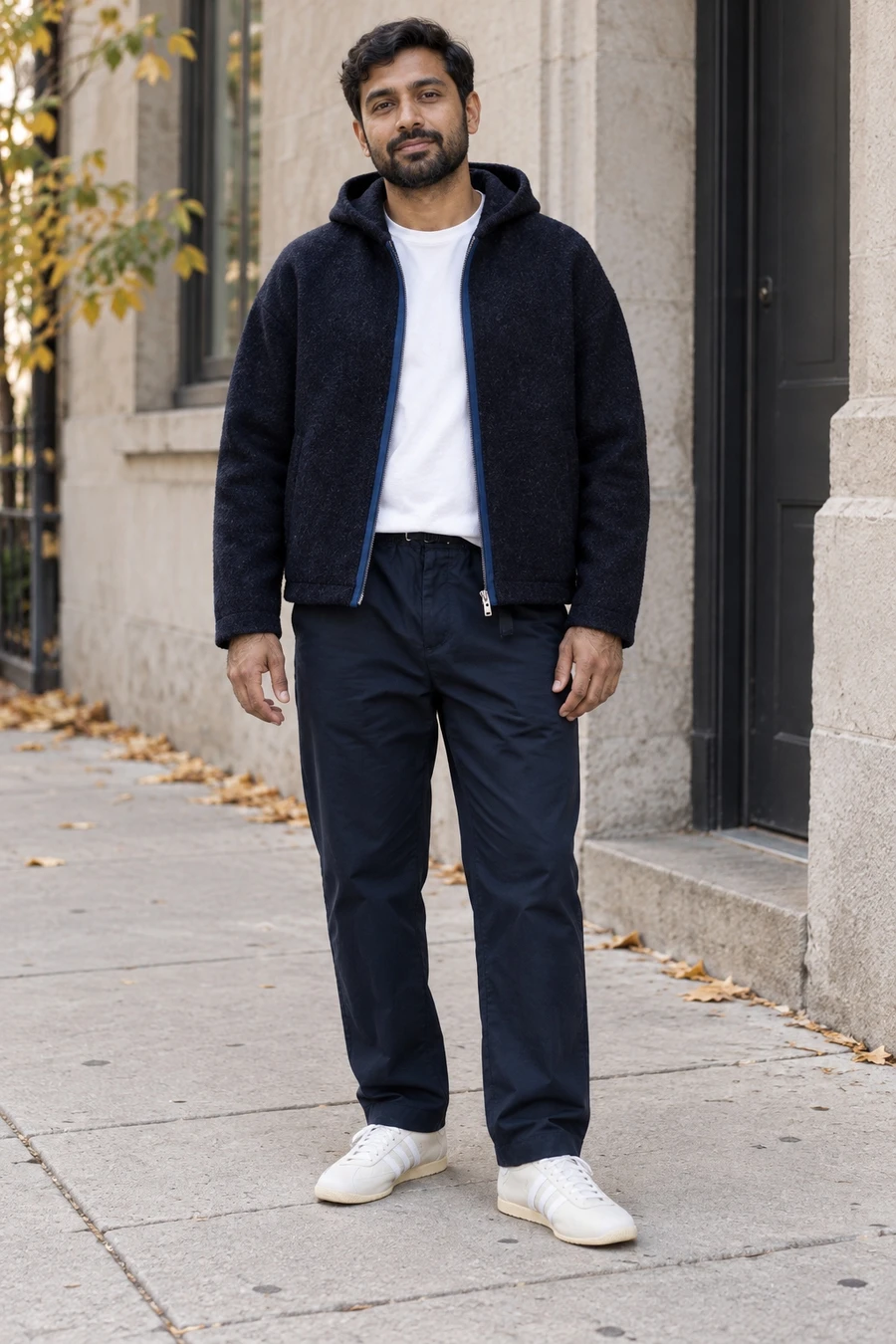 An adult man wears a navy hooded jacket, white tee and navy trousers with pale trainers on a Toronto sidewalk.