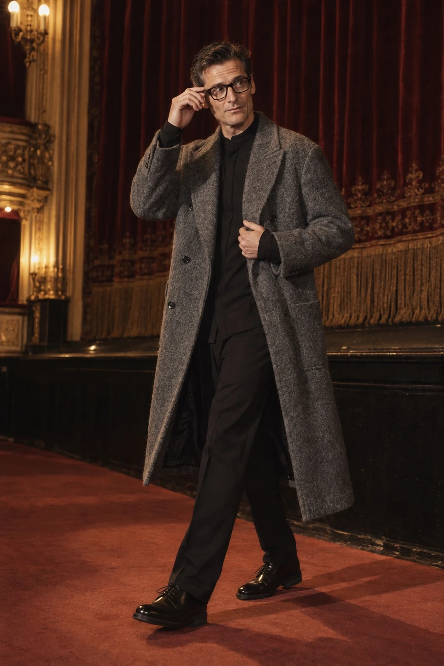 An English man in a grey COOR coat, a black thom/krom shirt, Avavav trousers, Louis Vuitton derbies and Chrome Hearts glasses stands before an opera house curtain in Vienna.