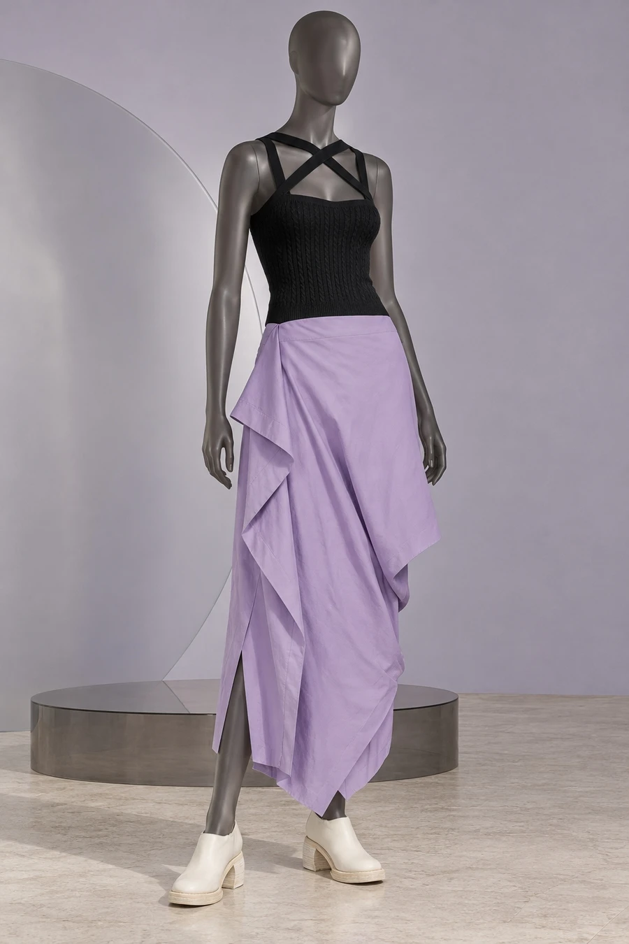 Black Patou crossed strap tank, lavender folded Issey Miyake skirt and ivory shoes on a dark frosted display.