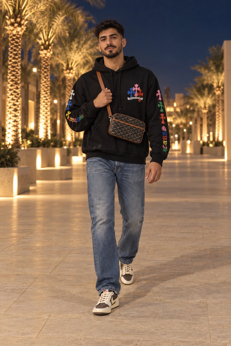 A young Saudi man in a black Chrome Hearts cross hoodie, Gallery Dept. jeans and Travis Scott Jordan 1 Lows walks across a lit Riyadh plaza at night with a Goyard crossbody.