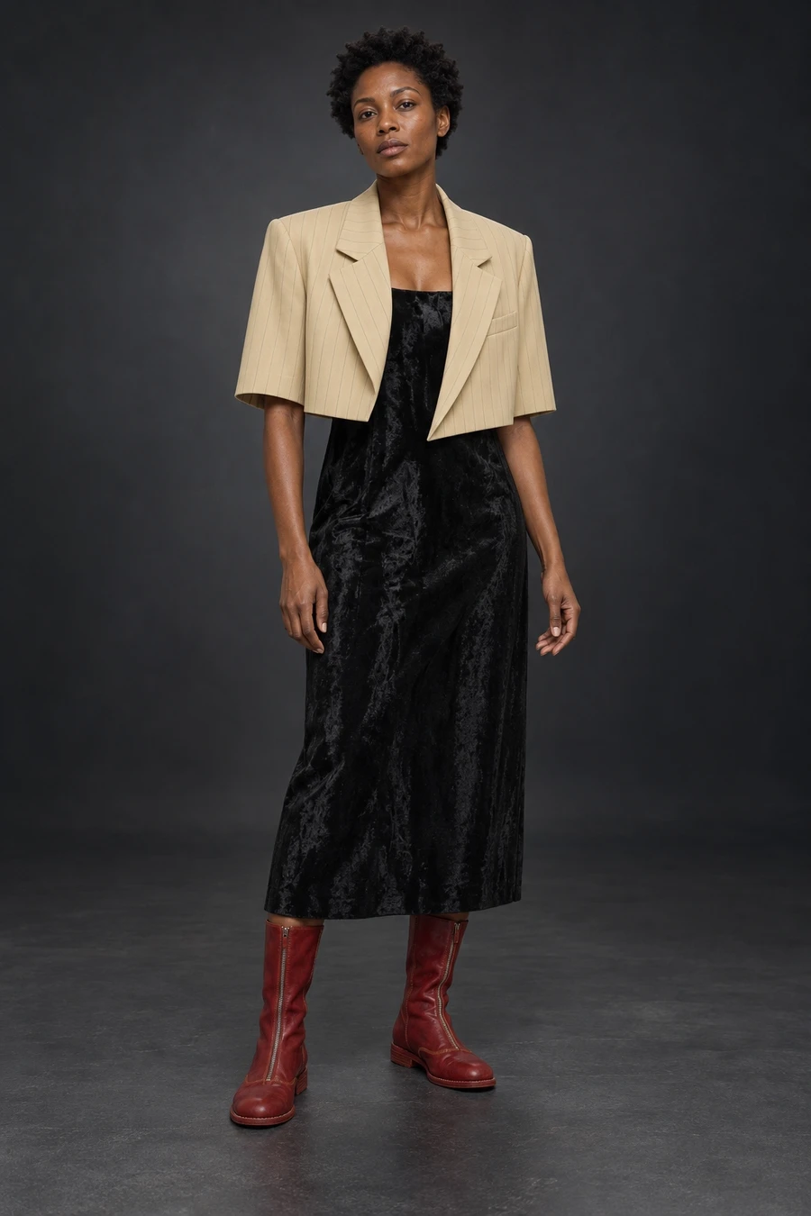 A cropped UJOH pinstripe jacket meets a black Helmut Lang midi dress and deep red Guidi boots. A woman stands against a dark studio background.