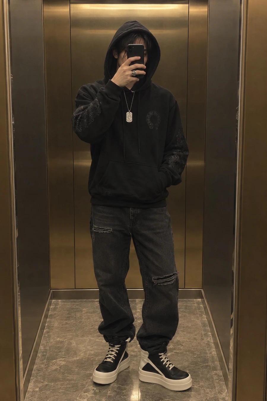 A Japanese man photographs his reflection in a brass lift mirror, wearing a black Chrome Hearts hoodie, washed black Represent jeans, Rick Owens Geobasket sneakers and a silver dog tag.