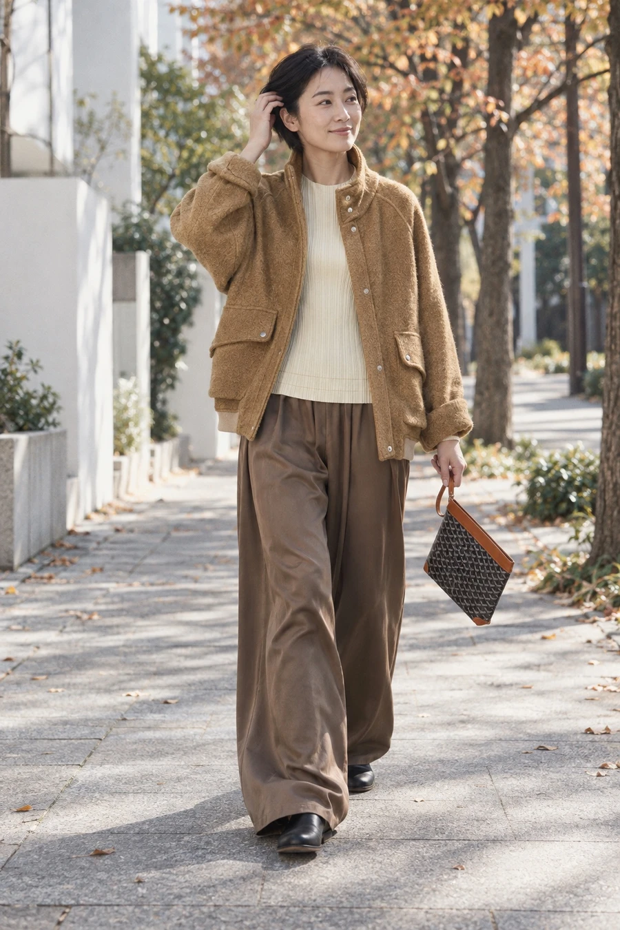 A Japanese woman in a camel Song for the Mute flight jacket, a pleated top, taupe RUOHAN silk trousers and black Guidi slip ons walks a Daikanyama street in Tokyo with a Goyard Conti pouch.